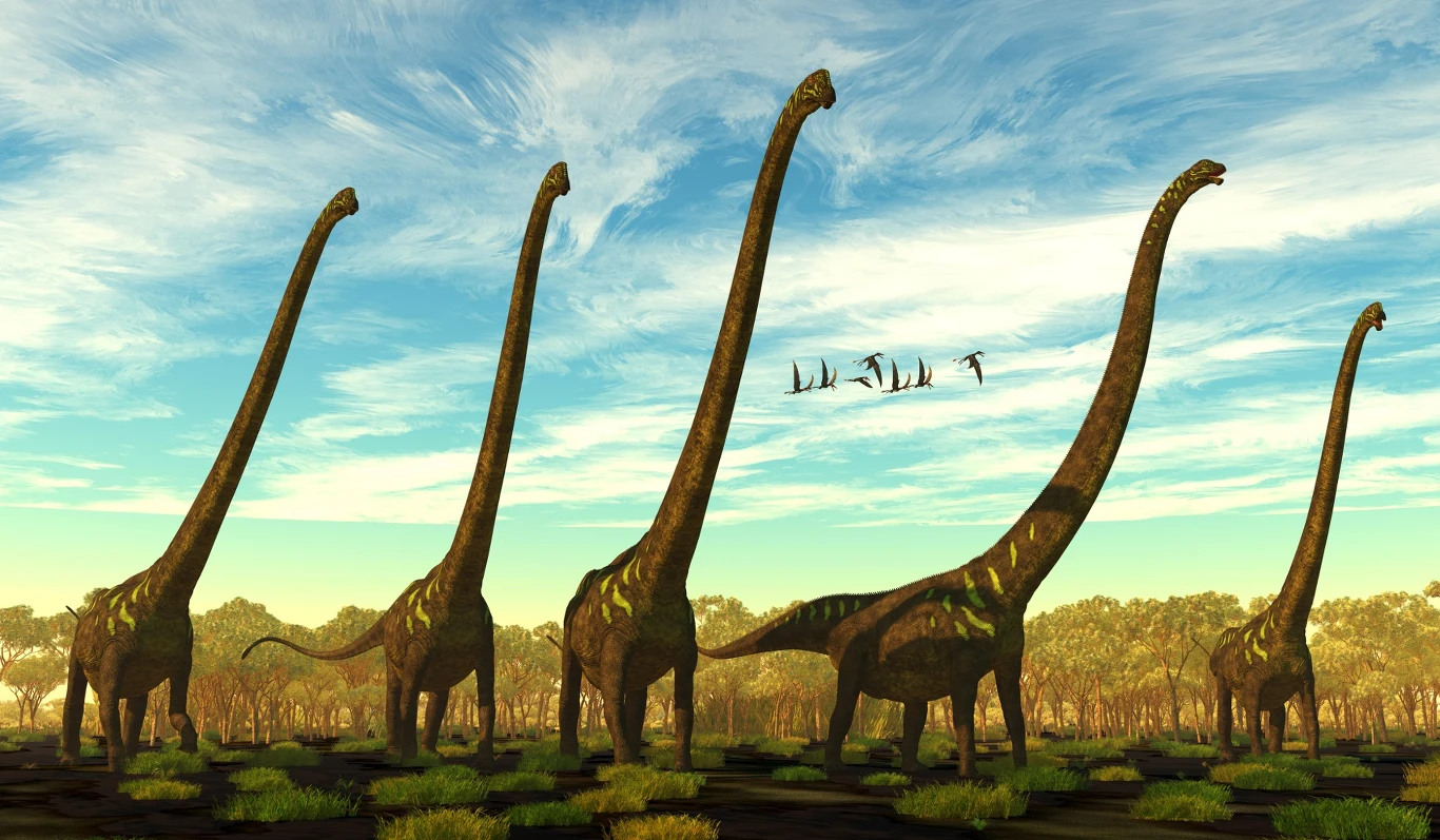 Mamenchizaury to jedne z największych dinozaurów. Znaleziono je w Chinach Stado długoszyich dinozaurów poruszających się po zielonym, prehistorycznym krajobrazie z niewielkimi kępami trawy i rozciągającym się w tle lasem, nad głowami przelatuje grupa ptaków na tle błękitnego, lekko zachmurzonego nieba.