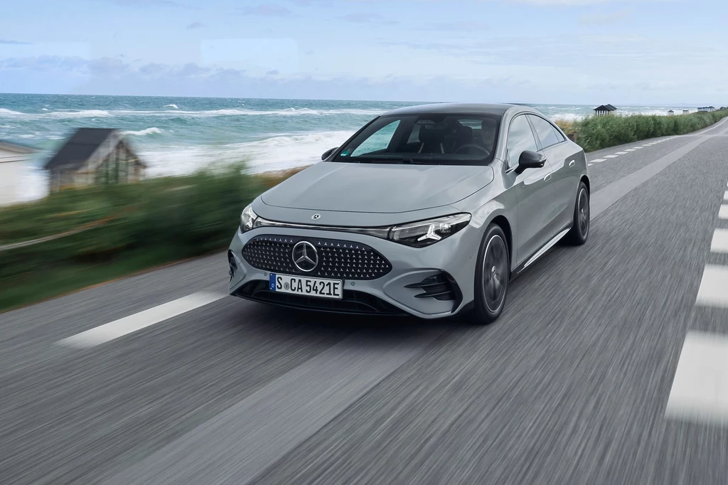 Podczas jazd testowych CLA 350 zużyło średnio 14,5 kWh na 100 km Srebrny samochód marki Mercedes poruszający się po asfaltowej drodze w pobliżu wybrzeża z widocznym morzem, zielenią i domkiem letniskowym na drugim planie.