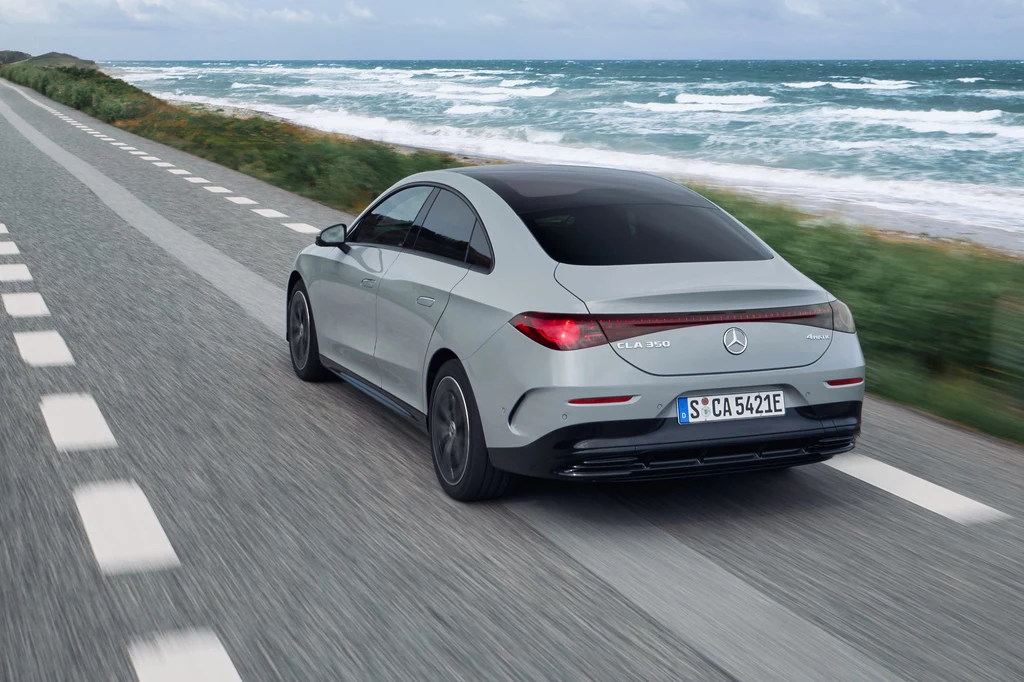 Nowe CLA imponuje poziomem wyciszenia wnętrza, nawet jak na elektryka Srebrny samochód marki Mercedes-Benz CLA 250e jedzie wzdłuż wybrzeża, po szerokiej, asfaltowej drodze, z widokiem na fale i linię brzegową po lewej stronie. Dynamiczny pejzaż, błękitne niebo, auto w ruchu.