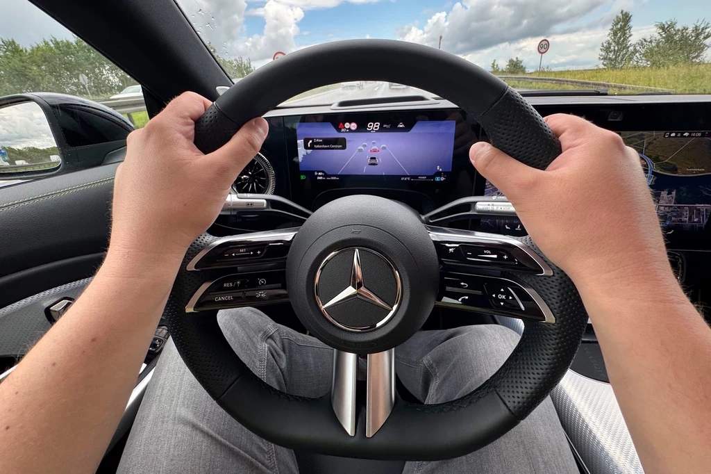 Na ekranie za kierownicą można obserwować cyfrową wersję otoczenia pojazdu Kierownica samochodu marki Mercedes trzymana oburącz przez kierowcę, cyfrowy kokpit wyświetlający mapę i prędkość, widok na drogę w tle przez przednią szybę.