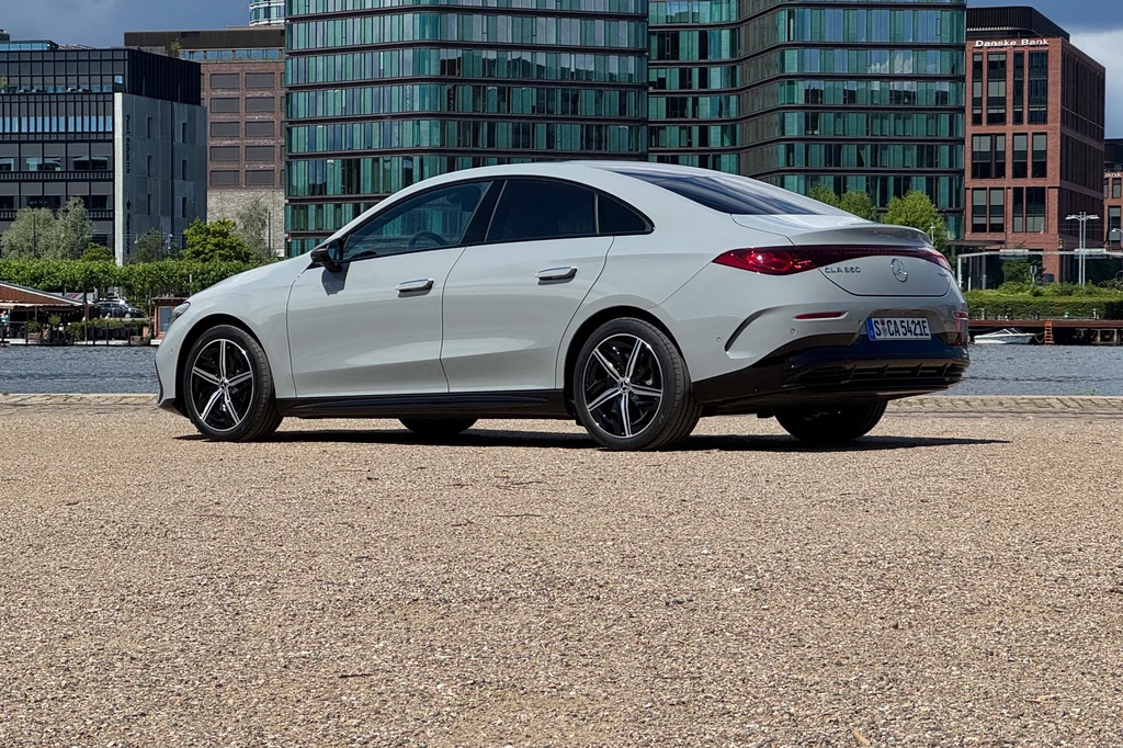 Elegancką linię nadwozia na postoju psują odstające klamki Biały samochód osobowy Mercedes CLA stojący na placu, w tle nowoczesne budynki biurowe ze szkła i betonu oraz zieleń miejskich drzew.