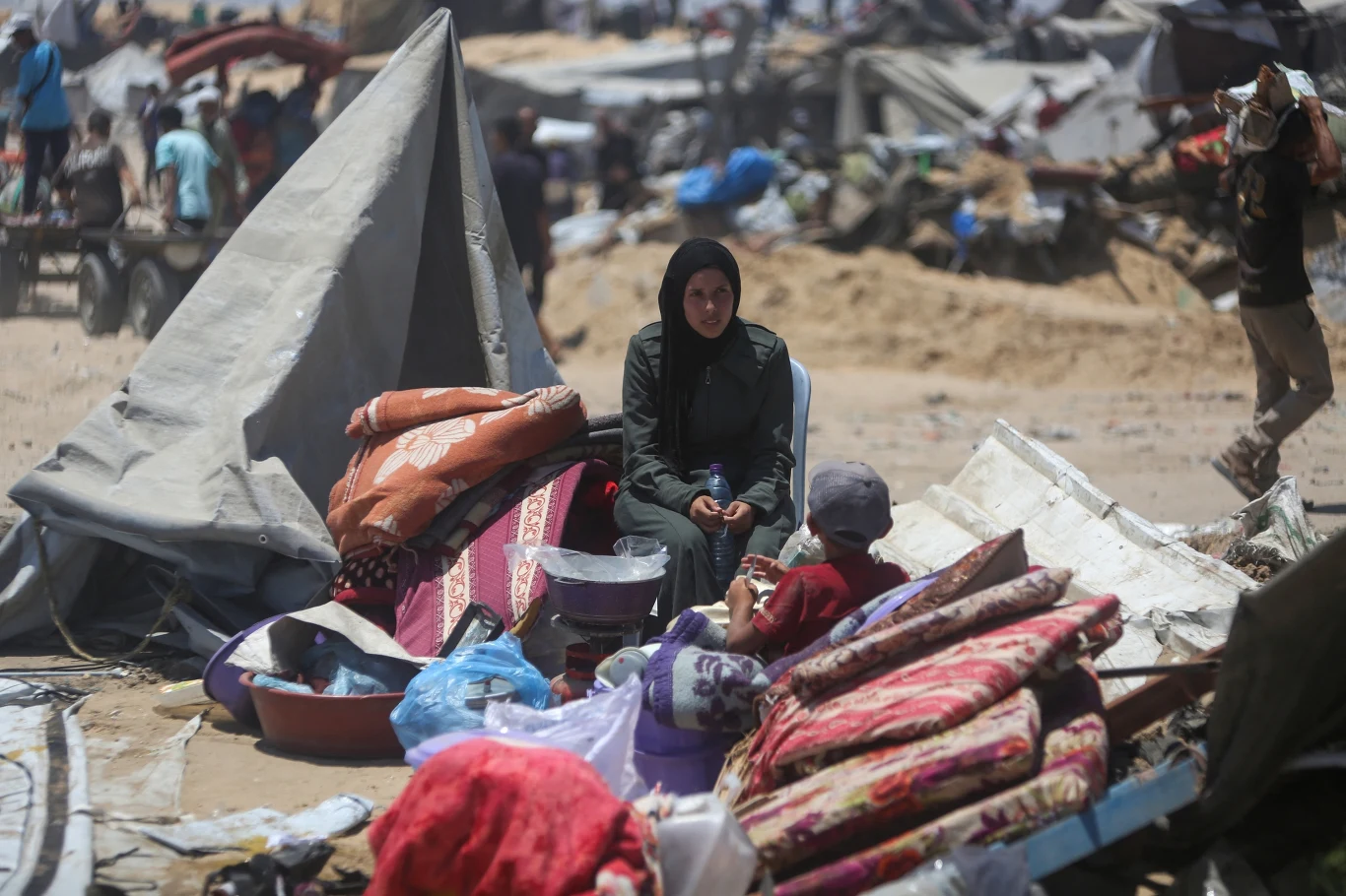 Kobieta w czarnym stroju siedzi wśród zniszczonych i porozrzucanych rzeczy, materacy oraz namiotów, obok niej dziecko, w tle widać innych ludzi i prowizoryczne schronienia na piaszczystym terenie.