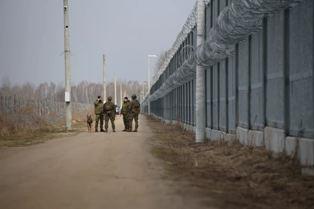Straż Graniczna patroluje okolice zapory na granicy polsko-białoruskiej Grupa żołnierzy z psem patroluje drogę biegnącą wzdłuż metalowego ogrodzenia z drutem kolczastym, po obu stronach widoczny teren otwarty oraz słupy oświetleniowe.