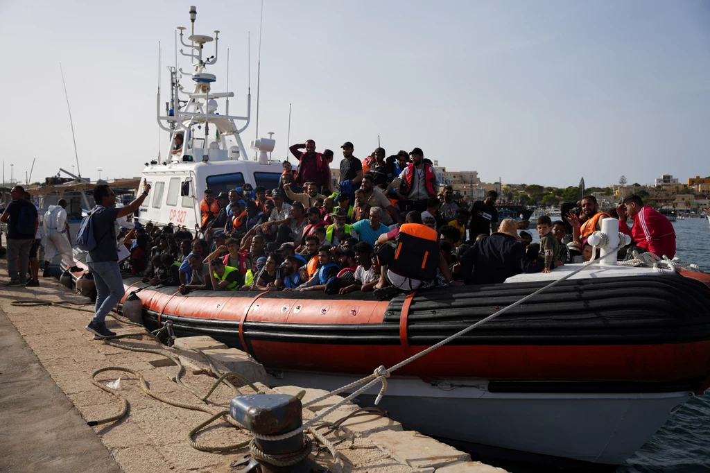 Migranci cumujący u wybrzeży włoskiej wyspy Lampedusa Duża grupa ludzi w kamizelkach ratunkowych siedzi na nadmuchiwanej łodzi przycumowanej do nabrzeża, w tle widoczny jest statek patrolowy oraz port.