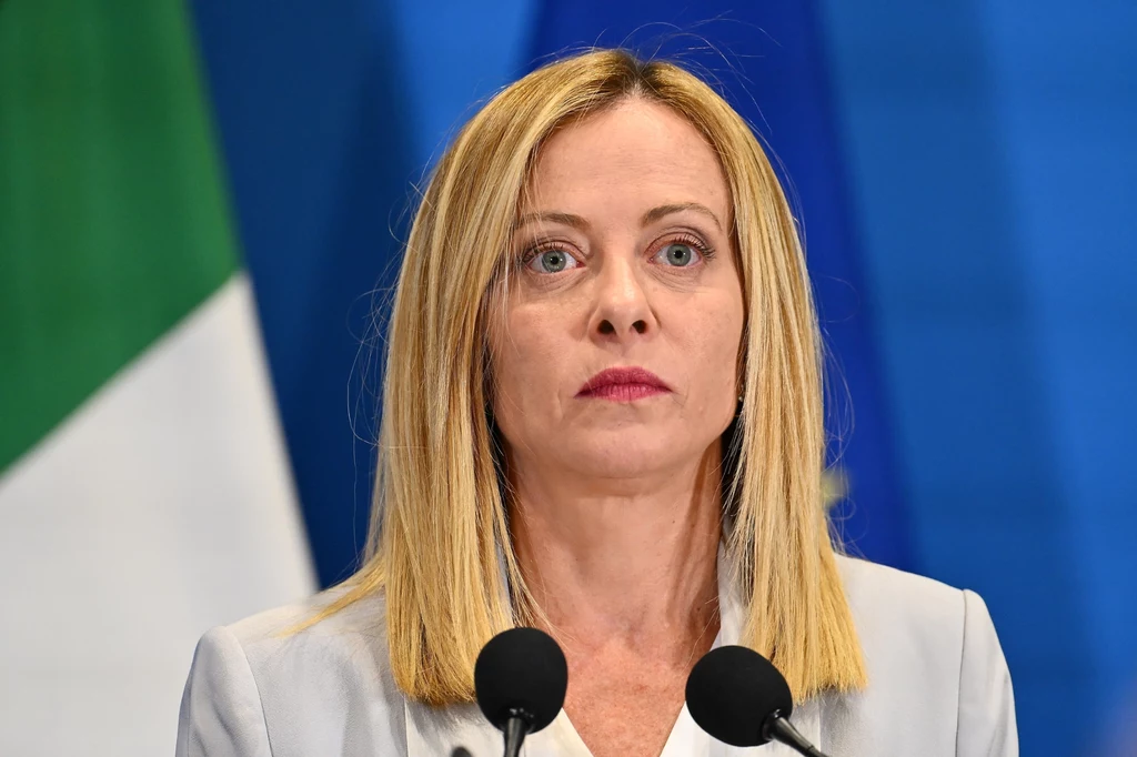 Premier Włoch Giorgia Meloni Kobieta o blond włosach w jasnej marynarce stoi przed mikrofonami, w tle fragment flagi włoskiej oraz niebieskie tło.