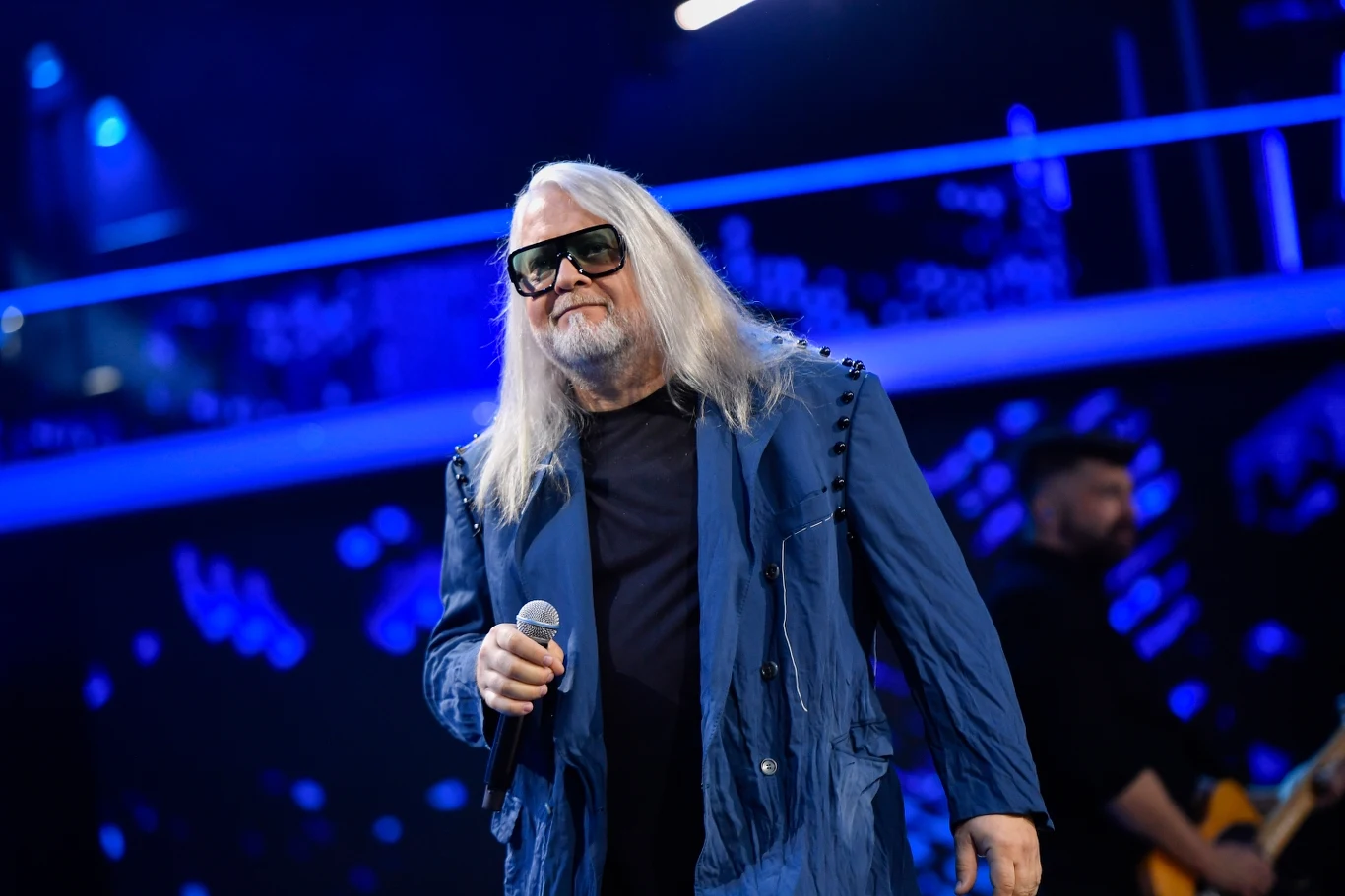 Artur Gadowski jest wokalistą grupy Ira Mężczyzna z długimi siwymi włosami i ciemnymi okularami trzyma mikrofon i stoi na scenie w niebieskim oświetleniu, w tle widoczny gitarzysta.