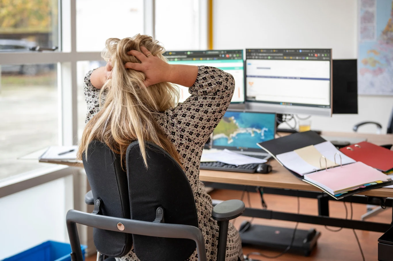 Kobieta siedząca przy biurku przed dwoma monitorami, trzymająca ręce na głowie w geście stresu, wokół niej rozłożone dokumenty i segregatory.