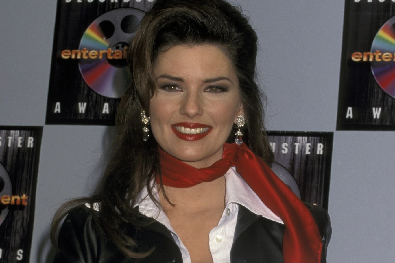 Shania Twain w latach 90. Uśmiechnięta kobieta ubrana w elegancki czarno-biały strój z czerwoną apaszką, stojąca na tle ściany z logotypami i napisem awards. Lysujące kolczyki i wyrazisty makijaż podkreślają jej wygląd.