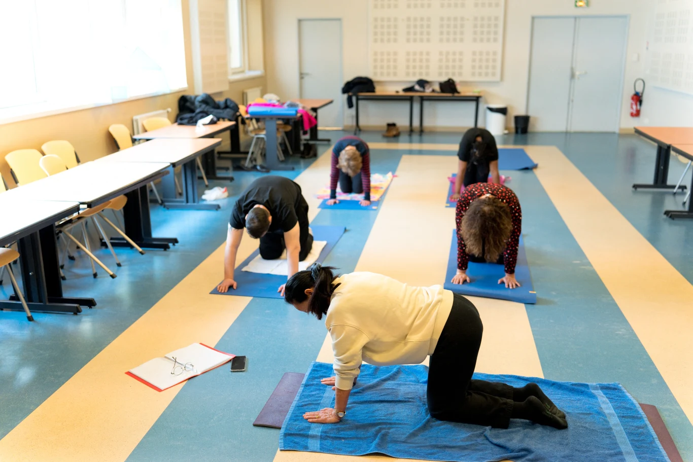 Grupa osób wykonuje ćwiczenia fizyczne na matach w sali lekcyjnej, wokół ustawione są stoły, krzesła oraz notatki i telefon, światło dzienne wpada przez duże okno po lewej stronie.