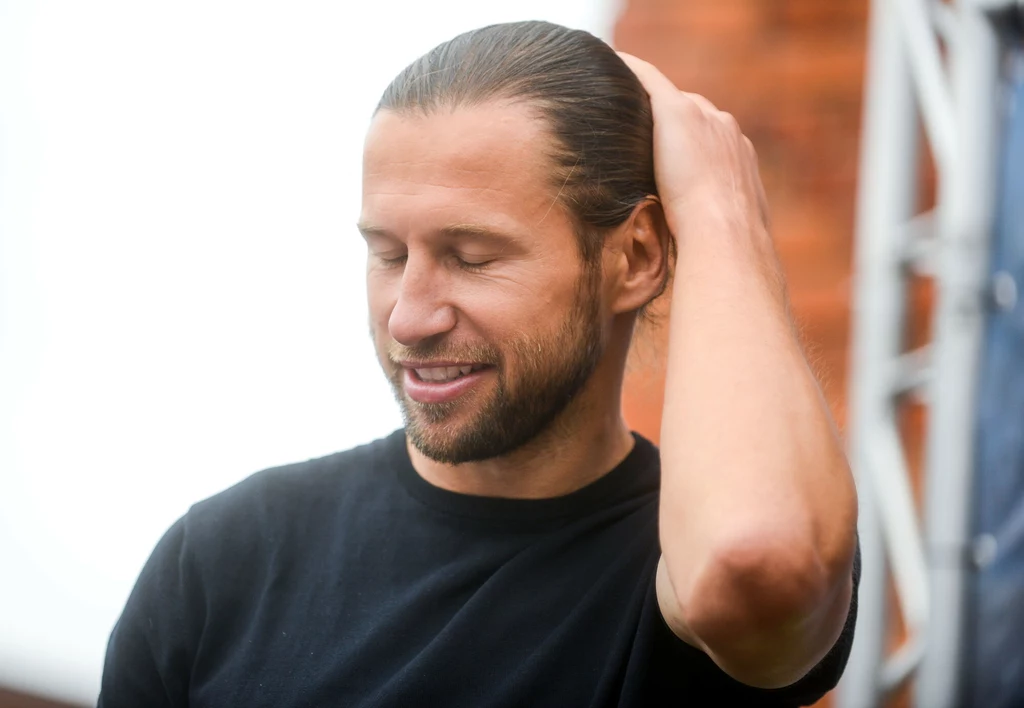 Grzegorz Krychowiak Mężczyzna z długimi włosami spiętymi do tyłu, ubrany w czarną koszulkę, trzyma rękę na głowie i delikatnie się uśmiecha, tło lekko rozmyte z konstrukcją metalową i ceglaną ścianą.