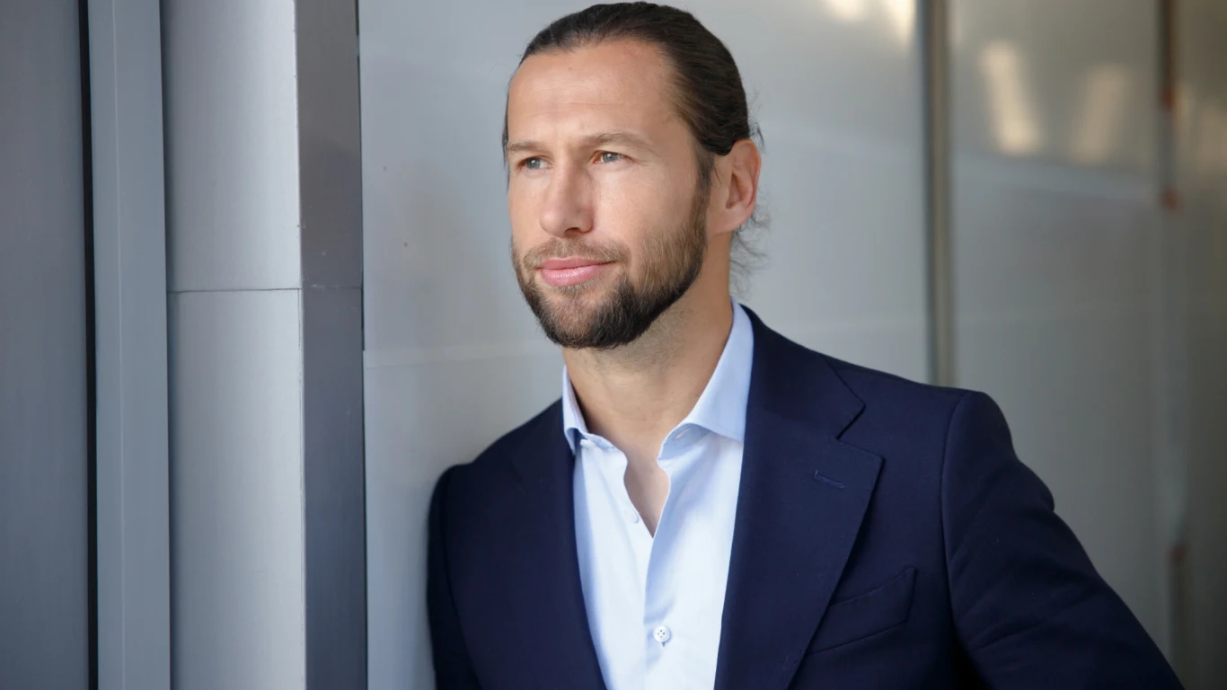 Mężczyzna z brodą i długimi włosami spiętymi w kucyk, ubrany w granatową marynarkę i jasnoniebieską koszulę, patrzy w dal z lekkim uśmiechem, oparty o ścianę w nowoczesnym, jasnym wnętrzu.