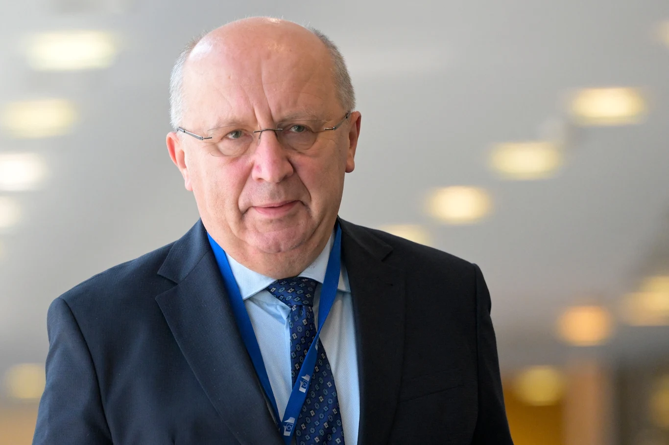 Andrius Kubilius proponuje budowę "muru dronowego" Starszy mężczyzna w okularach ubrany w granatowy garnitur, jasnoniebieską koszulę oraz krawat z wzorem, na szyi widoczna smycz z identyfikatorem, tło rozmyte z punktowymi światłami.