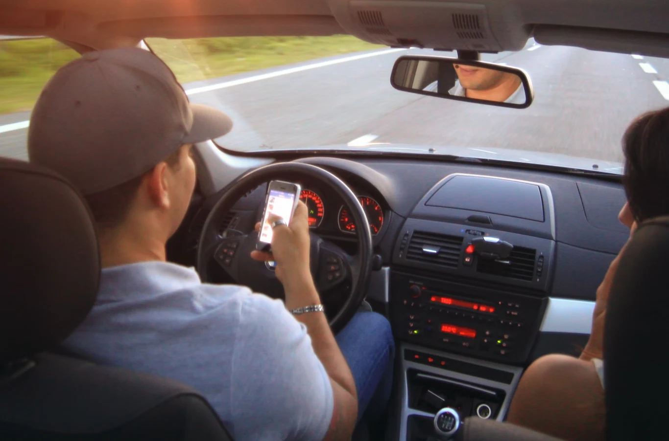 Nowy fotoradar Monocam wykrywa telefon w ręce kierowcy. Młody mężczyzna prowadzi samochód, jednocześnie trzymając w ręce telefon komórkowy i skupiając uwagę na ekranie, obok niego siedzi pasażerka, a pojazd jedzie po dwupasmowej drodze.