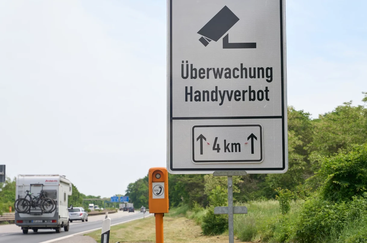 Monocam zarejestrował nawet 20 naruszeń na godzinę. Znaki drogowe informujące o monitoringu i zakazie korzystania z telefonu na odcinku 4 km, w tle autostrada z jadącymi samochodami i widoczna przydrożna kamera.