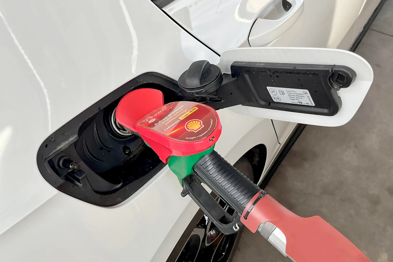 Eksperci przewidują obniżki cen paliw na stacjach Dystrybutor paliwa Shell z czerwonym pistoletem włożonym do wlewu paliwa białego samochodu, otwarta klapka wlewu i widoczne oznaczenia producenta na nakrętce zbiornika.