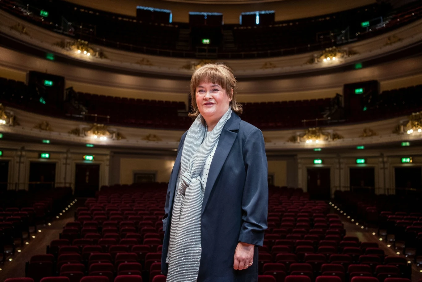Susan Boyle - zdobywczyni drugiego miejsca w trzeciej brytyjskiej edycji "Mam Talent" Kobieta w średnim wieku ubrana elegancko stoi na tle pustej widowni dużego teatru z balkonami oraz bogatą architekturą.