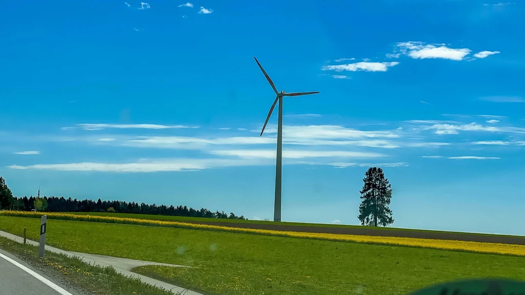 Niemcy postawią najwyższą na świecie turbinę wiatrową. To przełom w energetyce Pojedyncza turbina wiatrowa stoi na tle zielonego pola i niebieskiego, lekko zachmurzonego nieba, w pobliżu widoczna samotna sosna oraz fragment szosy.
