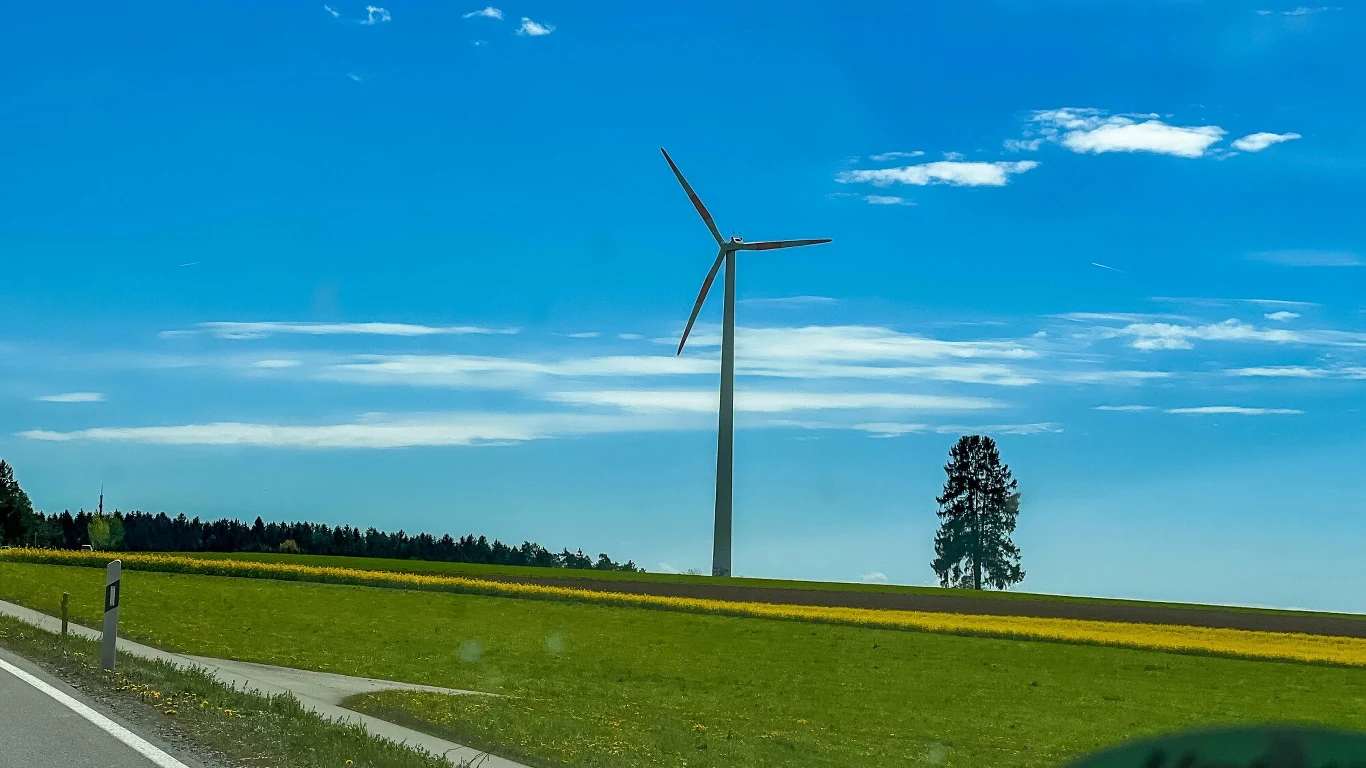 Niemcy postawią najwyższą na świecie turbinę wiatrową. To przełom w energetyce Pojedyncza turbina wiatrowa stoi na tle zielonego pola i niebieskiego, lekko zachmurzonego nieba, w pobliżu widoczna samotna sosna oraz fragment szosy.