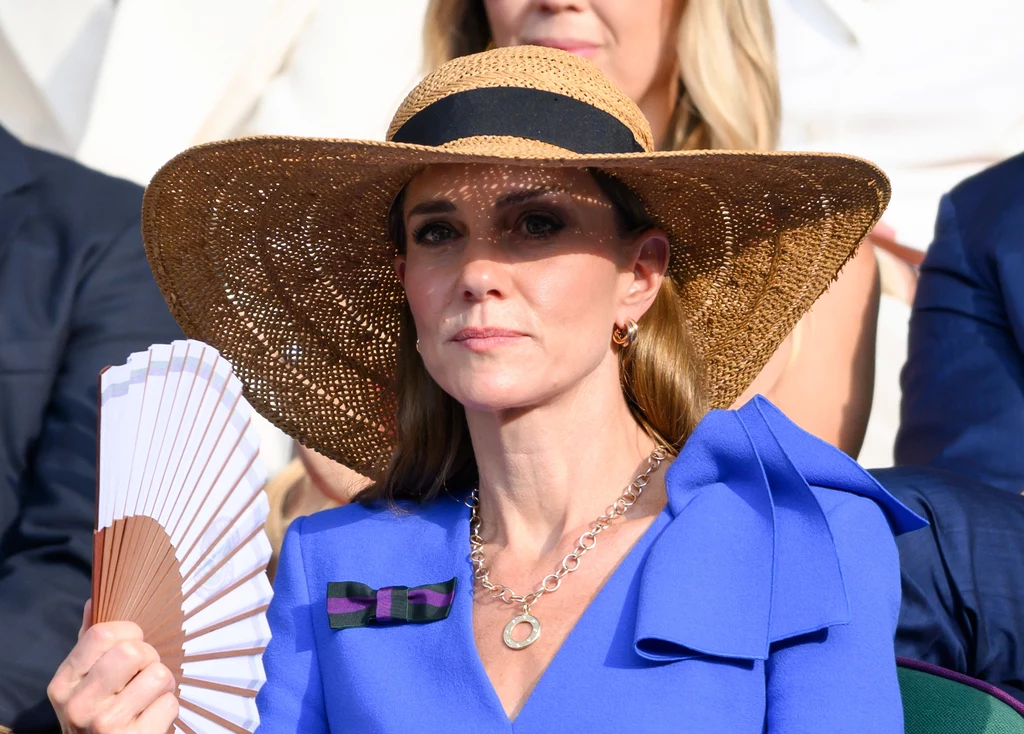 Księżna Kate wybierając kapelusz z szerokim rondem, dodała sobie kobiecości i tajemniczości Kobieta w eleganckim kapeluszu z szerokim rondem i niebieskim żakiecie siedzi na trybunach, trzyma wachlarz w ręce, ma na szyi srebrny naszyjnik i broszkę, w tle nieostre postaci innych widzów.
