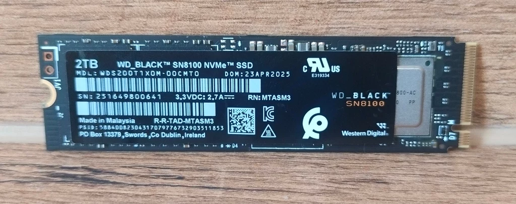 WD_Black SN8100 2TB SSD Pojemny dysk SSD WD_BLACK SN8100 NVMe o pojemności 2TB w standardzie M.2, czarna płytka drukowana z wyraźnymi oznaczeniami modelu, złączy oraz danych technicznych, całość leży na drewnianym blacie.