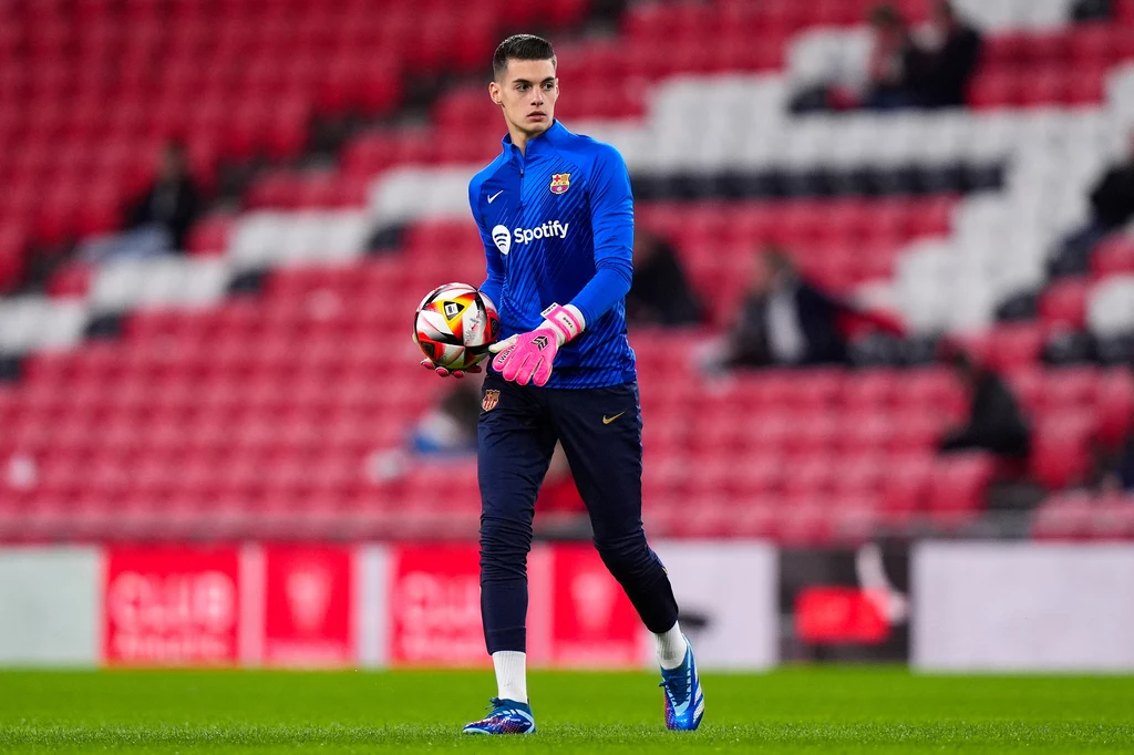 Aron Yaakobishvili Młody bramkarz w niebieskim dresie treningowym FC Barcelona trzyma piłkę w dłoniach, na stadionie z pustymi czerwonymi trybunami w tle.
