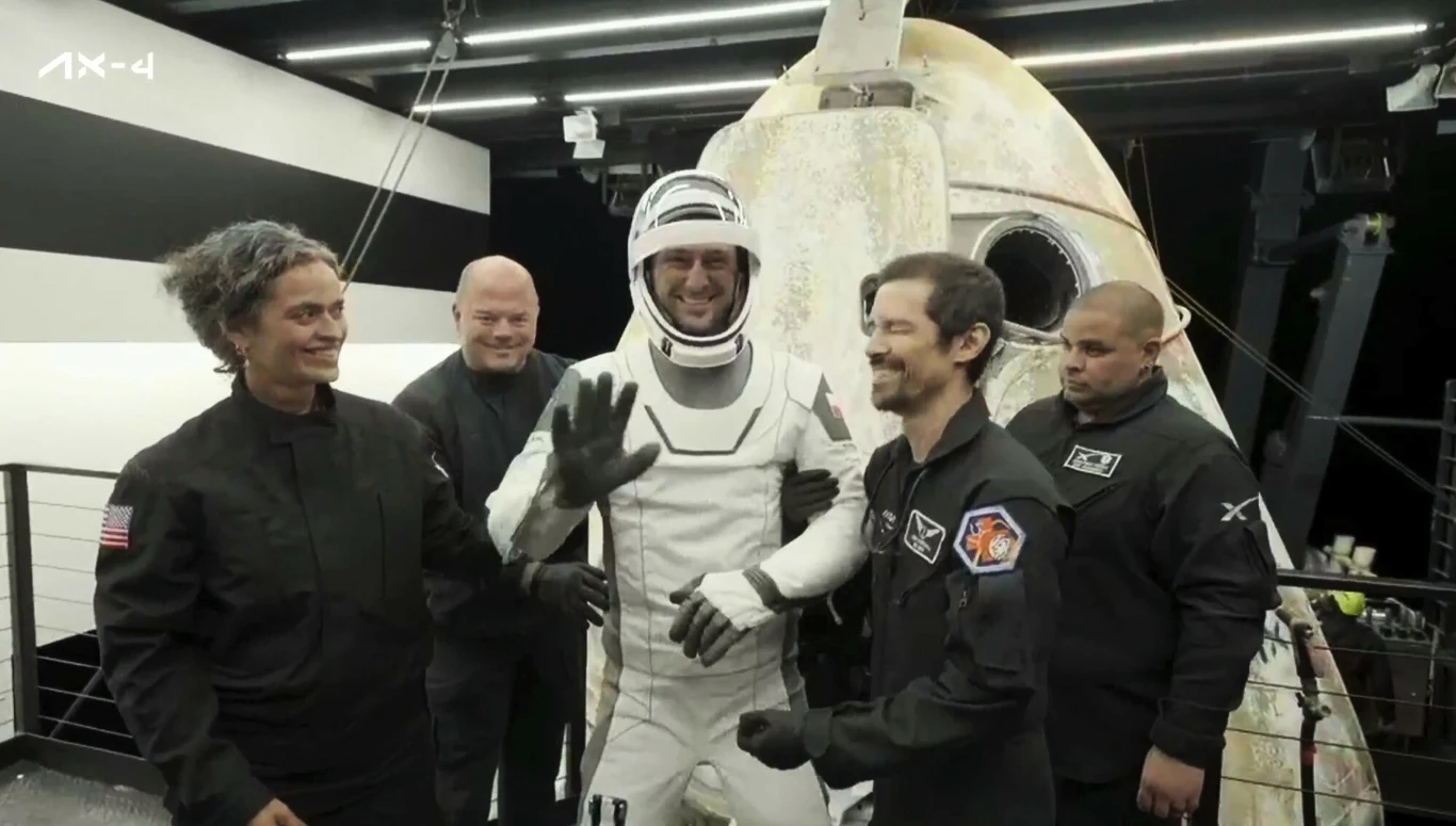 Grupa astronautów ubrana w kombinezony kosmiczne znajduje się przy kapsule kosmicznej, jeden z nich ubrany w biały skafander uśmiecha się i macha ręką, wokół panuje radosna atmosfera przygotowania lub powrotu z misji.