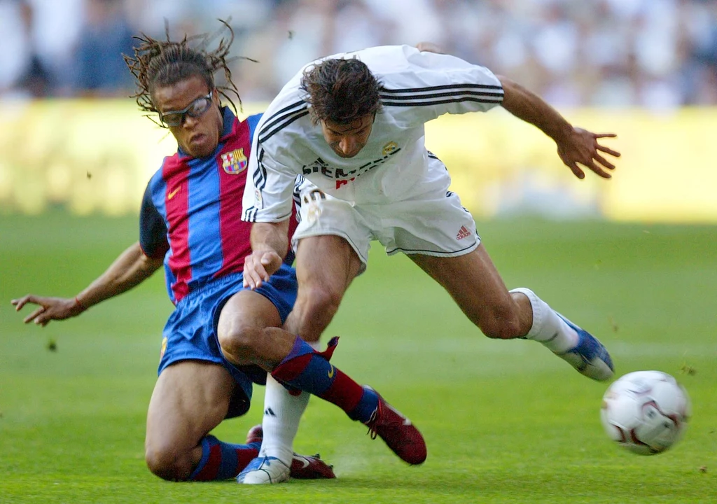 Edgar Davids w meczu Real Madryt - FC Barcelona. Walczy o piłkę z Luisem Figo Dwóch piłkarzy w dynamicznej walce o piłkę na boisku, zawodnik w białym stroju przechyla się tracąc równowagę, a drugi w kolorowej koszulce z wślizgiem próbuje odebrać piłkę.
