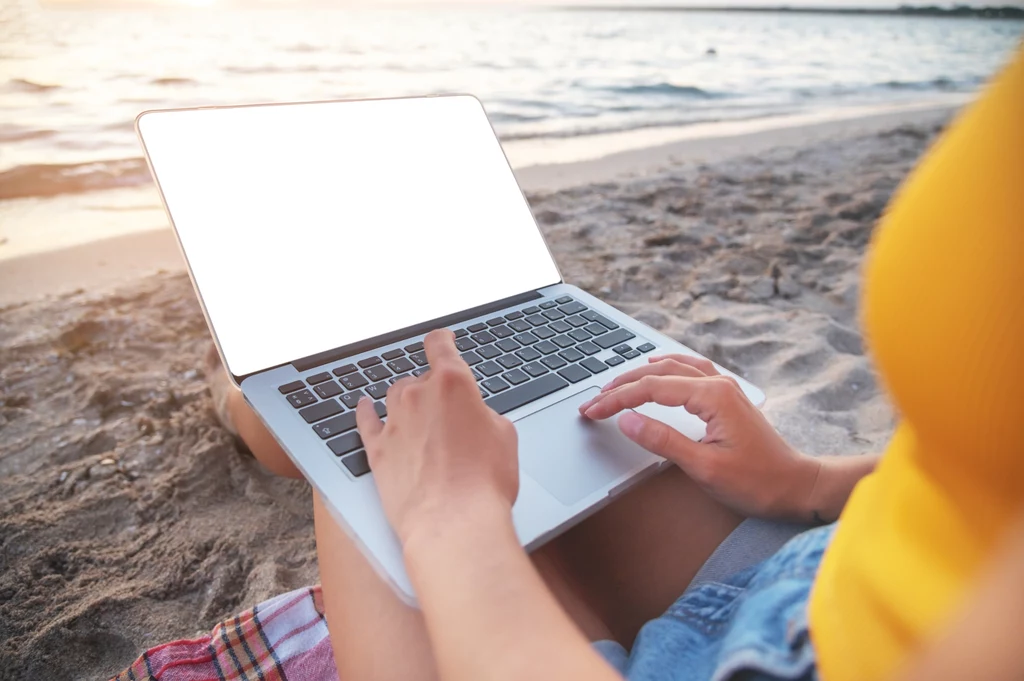Niektórzy nie mogą się powstrzymać przed odpisywaniem na maile, nawet na urlopie Osoba pracuje na laptopie na plaży, siedząc na piasku z widokiem na wodę. Jasny ekran komputera i letnia atmosfera podkreślają pracę zdalną lub relaks przy technologii nad morzem.