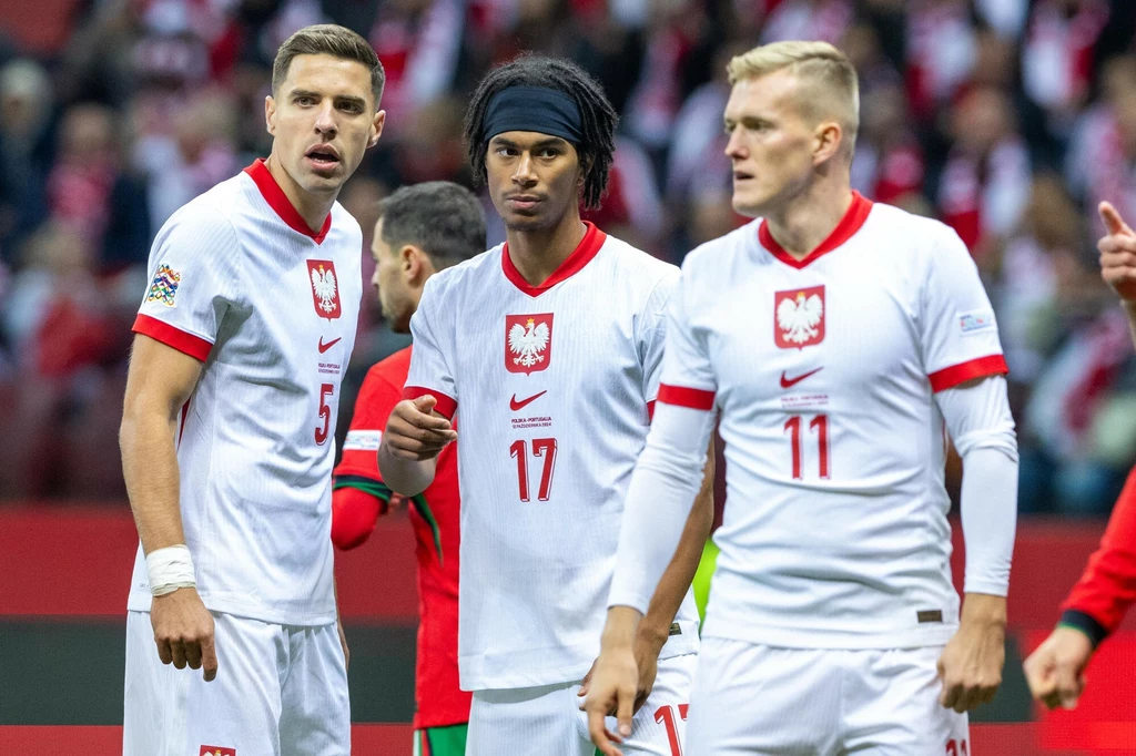 Jan Bednarek, Maxi Oyedele i Karol Świderski Trzech piłkarzy w białych strojach z orłem na piersi i czerwonymi akcentami stoi na murawie, skoncentrowani na grze, w tle widoczni kibice i zawodnicy drużyny przeciwnej w czerwonych strojach.