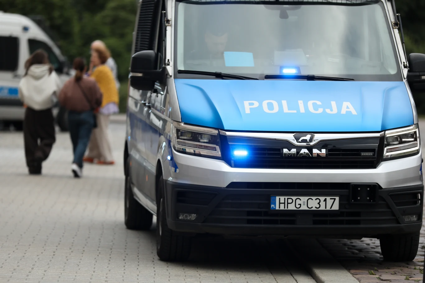 Dramatyczna zbrodnia na poznańskich Jeżycach (zdj. ilustracyjne) Radiowóz policyjny z włączonym niebieskim światłem stoi na ulicy, w tle kilka osób idących chodnikiem.