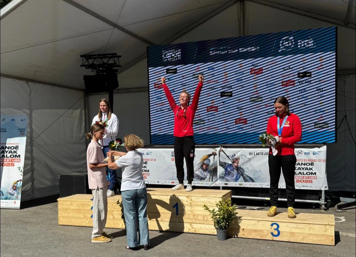 Ceremonia wręczenia nagród na podium podczas zawodów sportowych, trzy kobiety w sportowych strojach stoją na podium, środkowa zawodniczka unosi ręce w geście zwycięstwa, dwie osoby wręczają kwiaty laureatkom, w tle duży ekran z wynikami i baner zawodów.