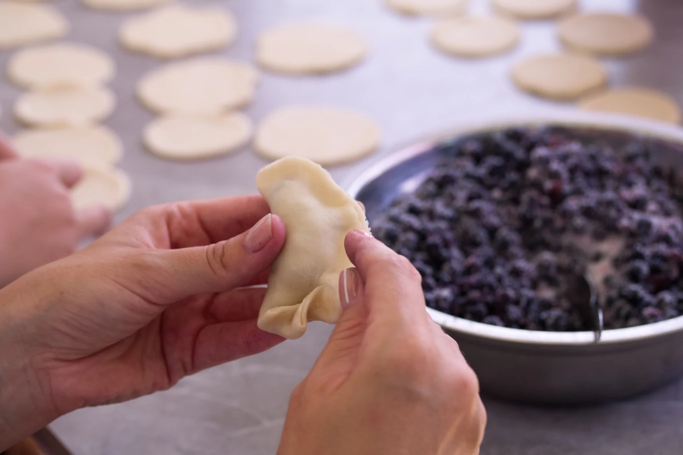 Ręce sklejające pieroga z ciasta nad stołem, na którym leży miska z nadzieniem z jagód oraz przygotowane krążki ciasta.