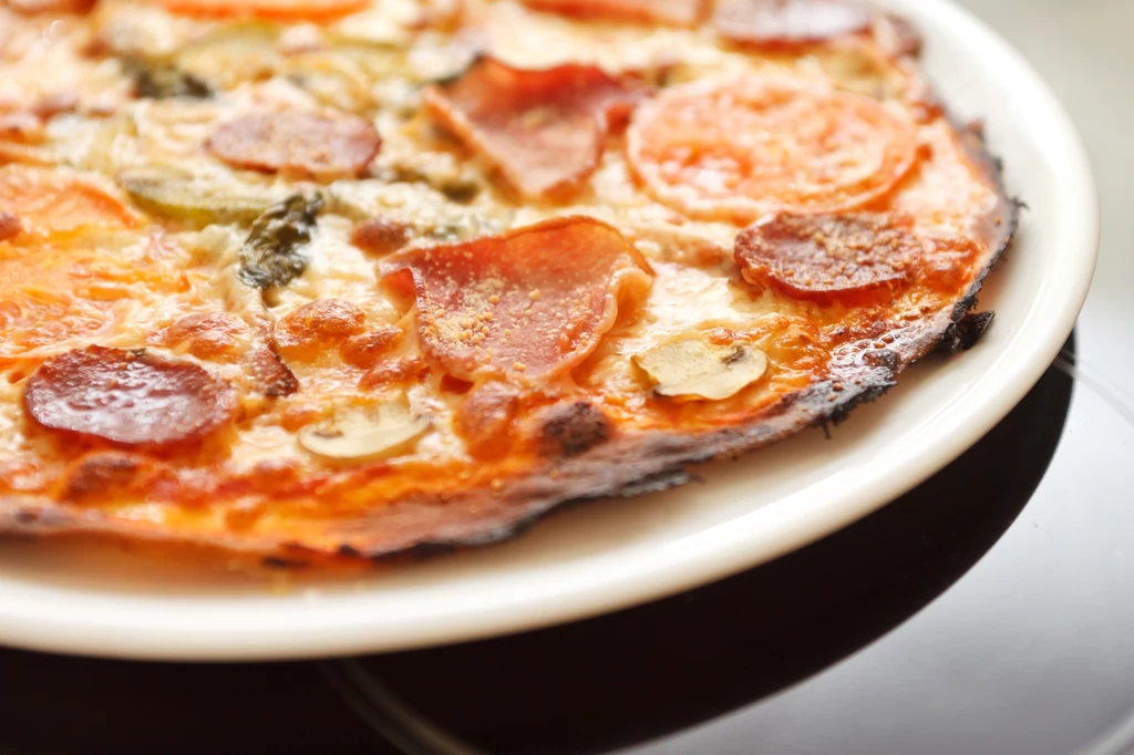 Pizza z cienkim ciastem, pokryta plasterkami salami, pomidorami, pieczarkami i ziołami, serwowane na białym talerzu.
