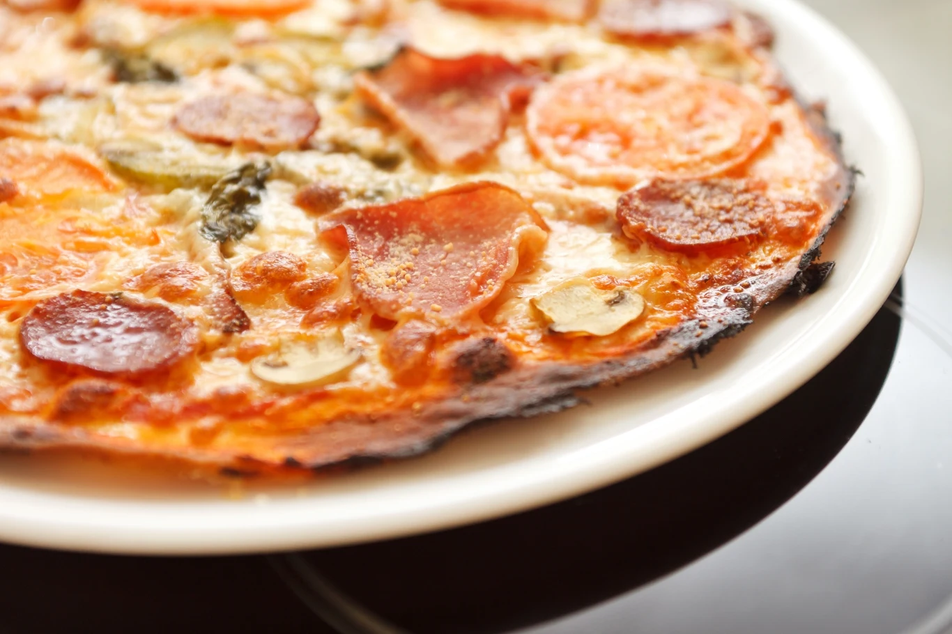 Pizza z cienkim ciastem, pokryta plasterkami salami, pomidorami, pieczarkami i ziołami, serwowane na białym talerzu.