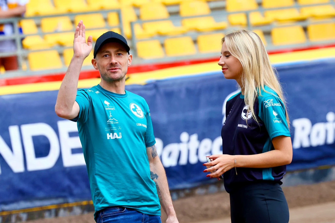 Mężczyzna w czapce z daszkiem unosi rękę w geście powitania, obok stoi uśmiechnięta kobieta ubrana w sportowy strój, w tle widać trybuny stadionu z żółtymi siedzeniami.