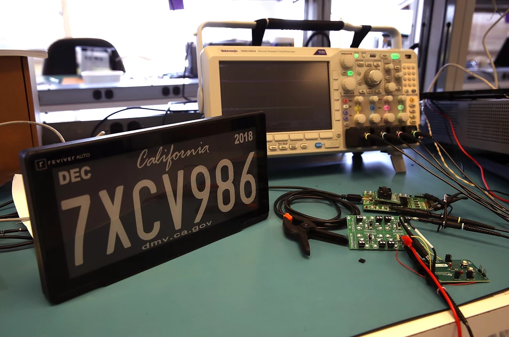 Cyfrowe posiadają dodatkowe pole do dynamicznego wyświetlania treści. Cyfrowa tablica rejestracyjna z napisem California 7XCV986 obok przyrządów elektronicznych, w tym oscyloskopu, kabli i płytek z elektroniką na biurku w laboratorium.