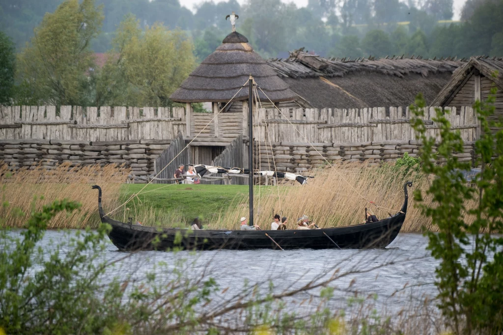 Drewniany okręt wikiński płynący po wodzie na tle rekonstrukcji grodu otoczonego palisadą, w oddali widać ludzi w historycznych strojach oraz tradycyjne chaty kryte strzechą.