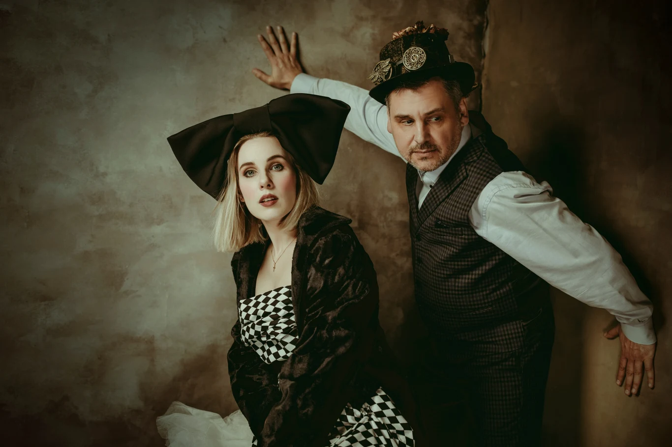 Dwie osoby w nietypowych stylizacjach pozują na tle ściany, kobieta w sukience w czarno-białą kratkę z dużą kokardą na głowie, mężczyzna w kamizelce i cylindrze ozdobionym kwiatami, oboje patrzą w innym kierunku, tworząc teatralny klimat.