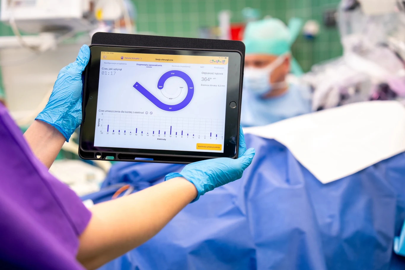 Tablet z wyświetlonym wykresem i danymi medycznymi trzymany przez osobę w rękawiczkach ochronnych nad pacjentem przykrytym niebieskim materiałem na sali operacyjnej, w tle zespół medyczny w maseczkach.
