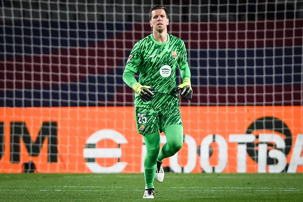 Wojciech Szczęsny Piłkarz w zielonym stroju bramkarza znajduje się na boisku piłkarskim, w tle widoczna jest siatka oraz wielkoformatowa reklama elektroniczna na stadionie.