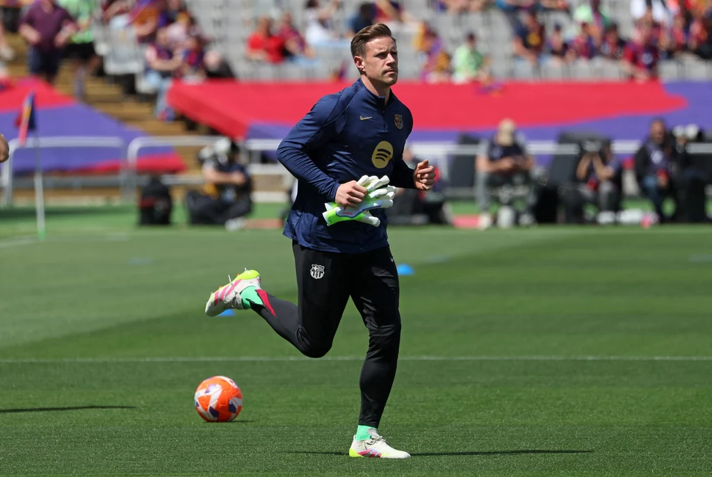 Marc-Andre ter Stegen Piłkarz w stroju sportowym biegnie po zielonej murawie stadionu, w tle widoczne są trybuny z kibicami oraz kolorowe banery.