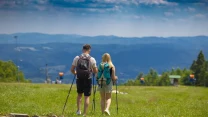Para turystów z plecakami i kijkami trekkingowymi spaceruje po zielonej łące, w tle widoczne są góry oraz lekko zamglony horyzont.
