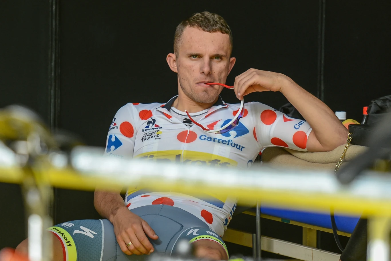 Kolarz w biało-czerwonej koszulce w grochy siedzi na ławce i żuje żel energetyczny, sprawiając wrażenie skoncentrowanego i zmęczonego po wyścigu rowerowym.