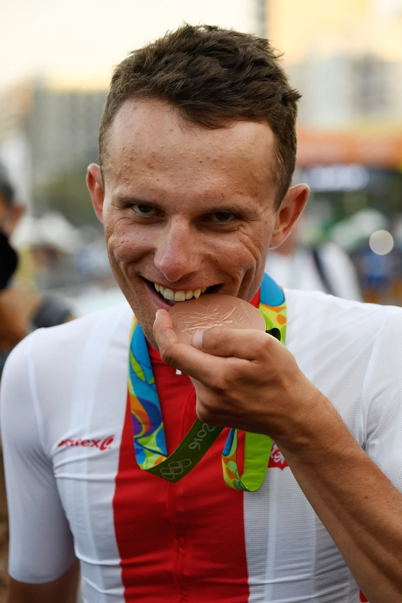 Sportowiec w biało-czerwonym stroju z medalem olimpijskim na szyi, trzyma medal blisko ust i uśmiecha się, wokół rozmyte sylwetki ludzi i miejskie tło.