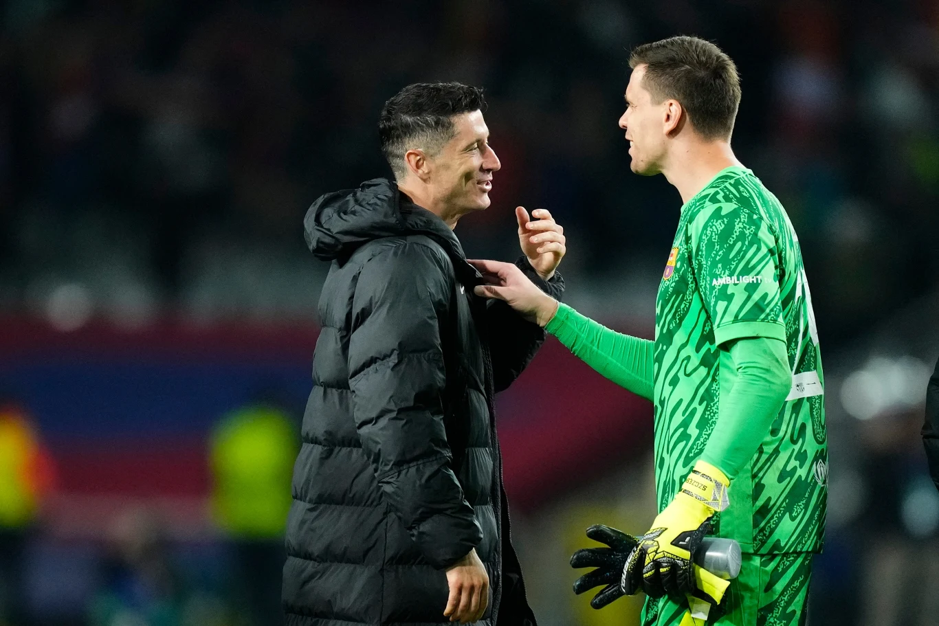 Rozmowa dwóch sportowców na boisku, jeden ubrany w długi czarny płaszcz, drugi w zielony strój bramkarski z rękawicami, w tle zamazana widownia stadionu.