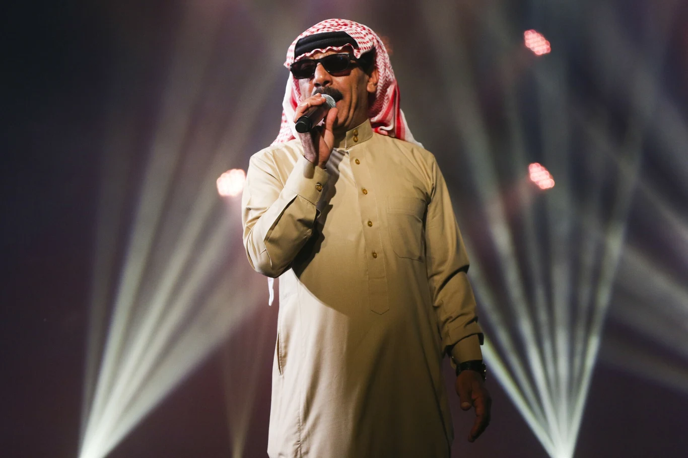Mężczyzna w tradycyjnym arabskim stroju i okularach przeciwsłonecznych śpiewa lub przemawia do mikrofonu na scenie, w tle intensywne światła reflektorów.