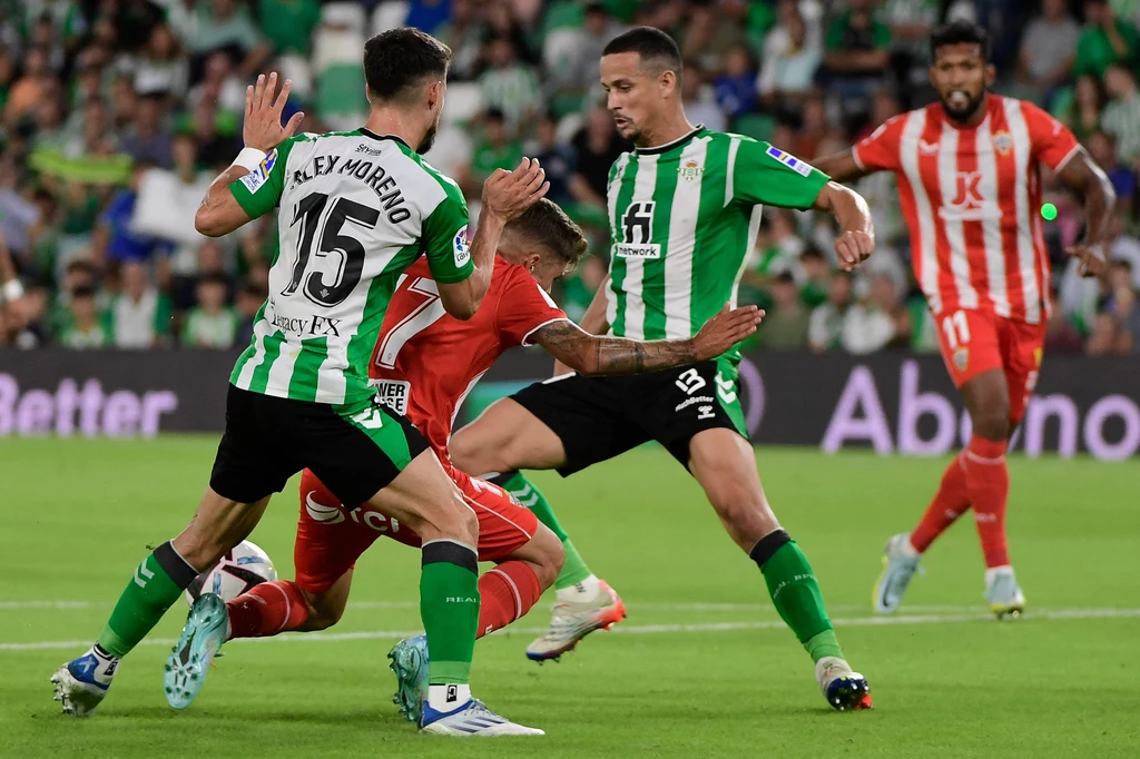 Alex Pozo kontra piłkarze Realu Betis Zawodnicy dwóch drużyn piłkarskich w zielono-białych i czerwonych strojach rywalizują o piłkę na murawie stadionu, w tle kibice obserwujący przebieg meczu.