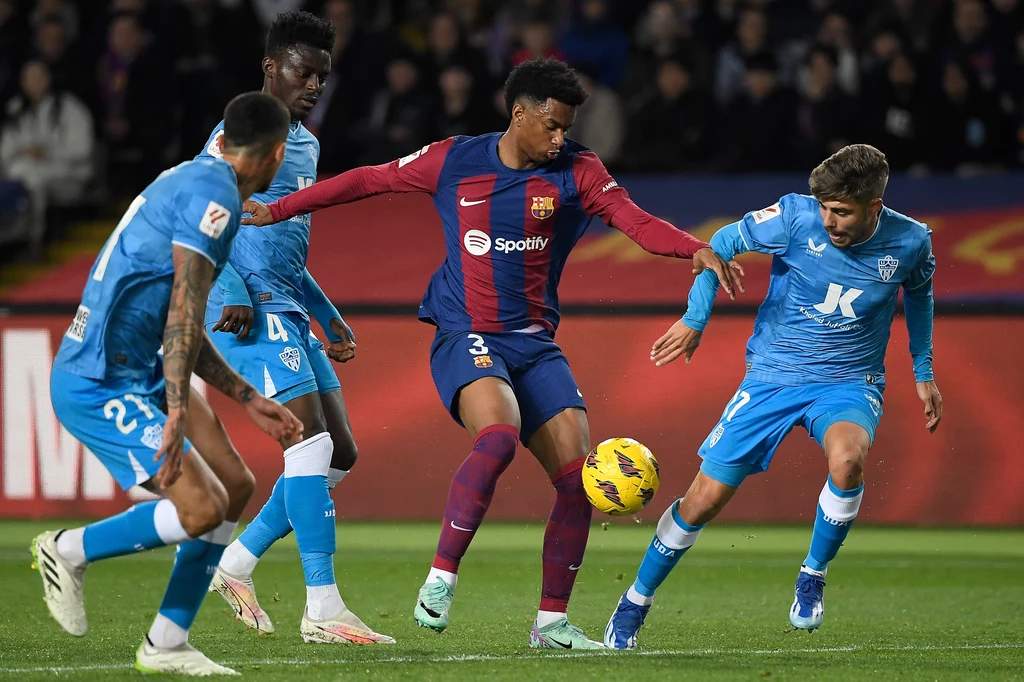 Alex Pozo (pierwszy z prawej) w meczu przeciwko FC Barcelona Czterech piłkarzy w trakcie dynamicznej walki o piłkę na boisku, w centrum gracz w bordowo-granatowym stroju otoczony przez zawodników drużyny w błękitnych koszulkach, w tle widoczna publiczność