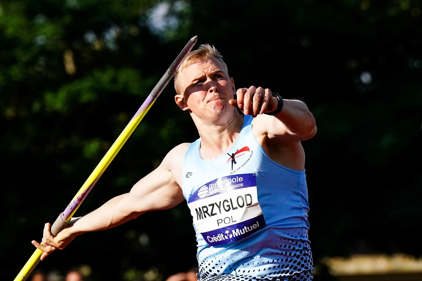 Cyprian Mrzygłód Mężczyzna w sportowym stroju podczas dynamicznego rzutu oszczepem na tle rozmytego, ciemnego otoczenia, widoczny numer startowy z napisem MRZYGŁÓD POL.
