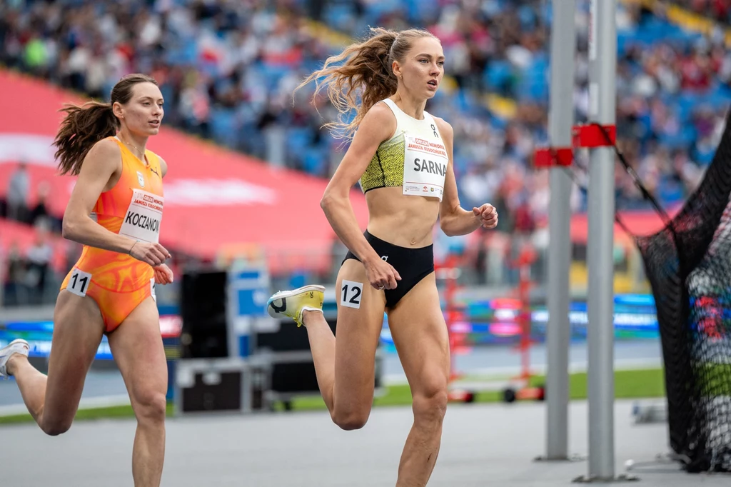 Angelika Sarna i Margarita Koczanowa Dwie biegaczki podczas wyścigu na stadionie, jedna z nich jest na prowadzeniu, w tle niewyraźna publiczność oraz elementy stadionu.